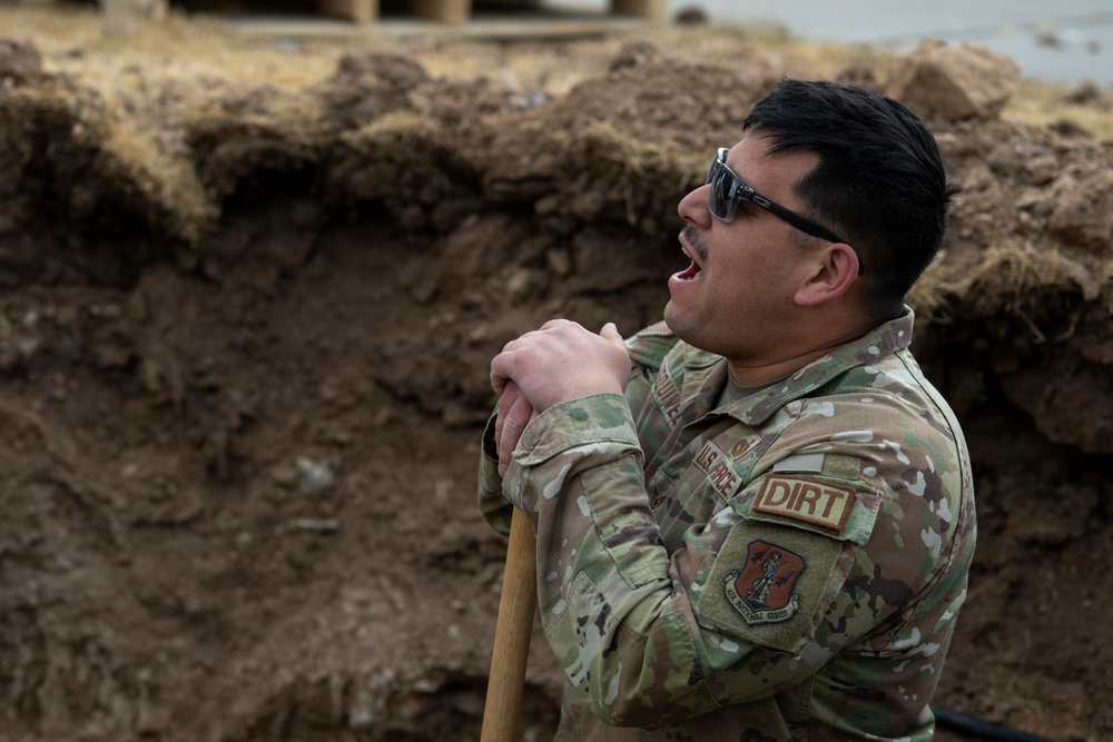 A U.S. Air Force National Guard service member is standing in or near an excavated trench, leaning on a shovel handle with mouth open, appearing to be speaking or calling out. He is wearing OCP (Operational Camouflage Pattern) uniform with visible patches including 'U.S. AIR FORCE', 'DIRT', and an Air National Guard shoulder patch. The setting appears to be an outdoor construction or field engineering work site.