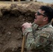 A U.S. Air Force National Guard service member is standing in or near an excavated trench, leaning on a shovel handle with mouth open, appearing to be speaking or calling out. He is wearing OCP (Operational Camouflage Pattern) uniform with visible patches including 'U.S. AIR FORCE', 'DIRT', and an Air National Guard shoulder patch. The setting appears to be an outdoor construction or field engineering work site.