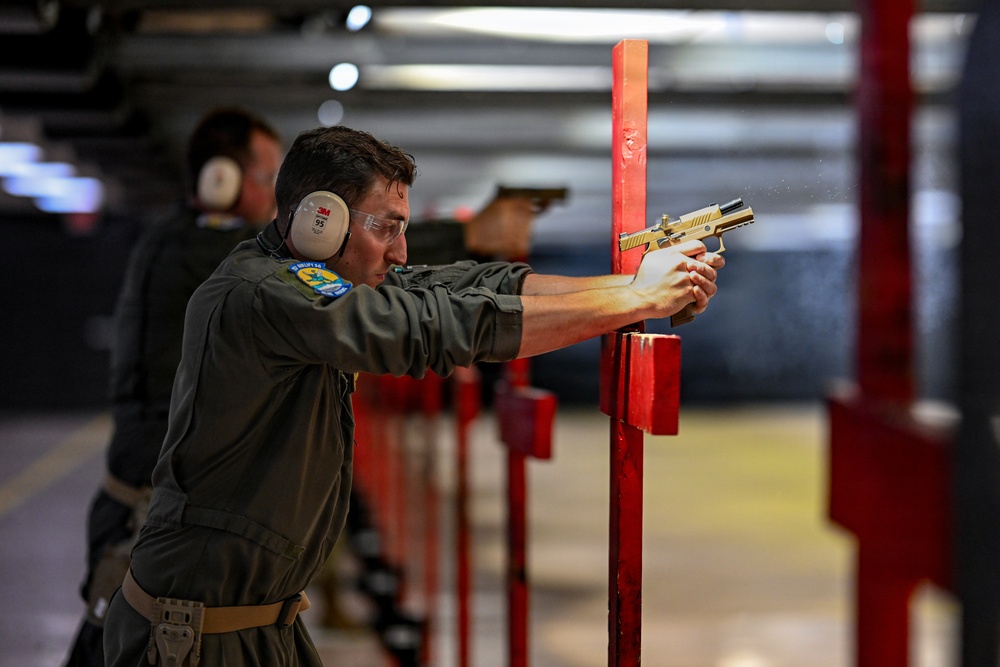 The image shows multiple individuals at an indoor shooting range, with the foreground subject in focus wearing olive/gray uniform, ear protection, and safety glasses while firing a gold/tan-colored semi-automatic pistol. The shooter is positioned at a red vertical divider post, using a two-handed grip stance. Additional shooters are visible in the background in similar positions along the range lane dividers.