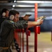 The image shows multiple individuals at an indoor shooting range, with the foreground subject in focus wearing olive/gray uniform, ear protection, and safety glasses while firing a gold/tan-colored semi-automatic pistol. The shooter is positioned at a red vertical divider post, using a two-handed grip stance. Additional shooters are visible in the background in similar positions along the range lane dividers.