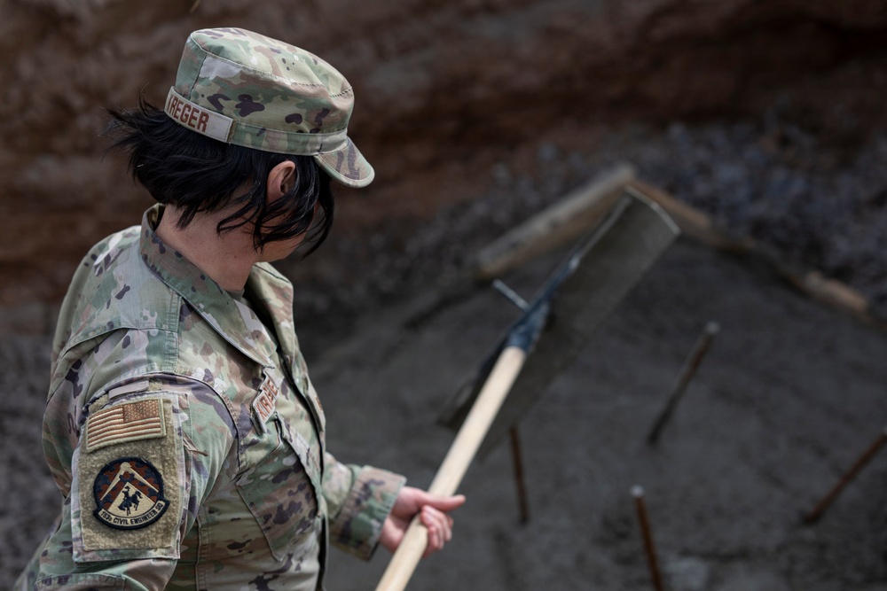A U.S. Air Force service member wearing Operational Camouflage Pattern (OCP) uniform is engaged in construction or engineering work at what appears to be a job site. The individual's name tape reads 'KREGER' and their sleeve patch indicates affiliation with the 807th Civil Engineer Squadron. They are holding a rake or similar hand tool over a gravel or aggregate surface with visible rebar.