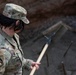 A U.S. Air Force service member wearing Operational Camouflage Pattern (OCP) uniform is engaged in construction or engineering work at what appears to be a job site. The individual's name tape reads 'KREGER' and their sleeve patch indicates affiliation with the 807th Civil Engineer Squadron. They are holding a rake or similar hand tool over a gravel or aggregate surface with visible rebar.
