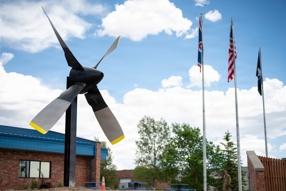 The image shows an outdoor display featuring a large aircraft propeller mounted on a post as a static exhibit, likely at a military or aviation facility. Three flagpoles are visible in the background flying what appear to be a state flag, the United States flag, and a POW/MIA flag. A brick building with a blue roof is visible in the background along with trees and a partly cloudy sky.