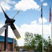 The image shows an outdoor display featuring a large aircraft propeller mounted on a post as a static exhibit, likely at a military or aviation facility. Three flagpoles are visible in the background flying what appear to be a state flag, the United States flag, and a POW/MIA flag. A brick building with a blue roof is visible in the background along with trees and a partly cloudy sky.