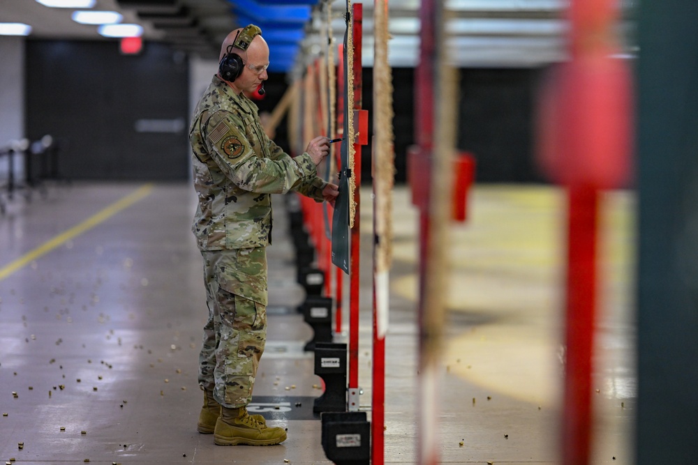 A military service member in OCP (Operational Camouflage Pattern) uniform is standing at an indoor shooting range, examining or scoring a target board. The floor is littered with spent brass casings, indicating recent firing activity. The range features multiple lanes with red divider posts and target retrieval systems.