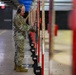 A military service member in OCP (Operational Camouflage Pattern) uniform is standing at an indoor shooting range, examining or scoring a target board. The floor is littered with spent brass casings, indicating recent firing activity. The range features multiple lanes with red divider posts and target retrieval systems.
