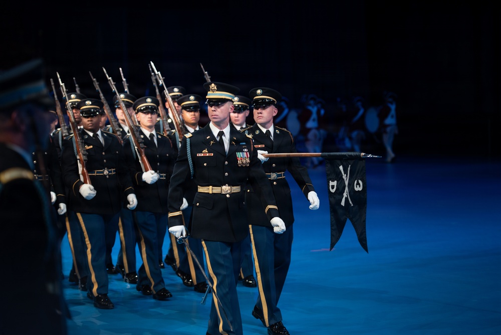 A formal military drill or ceremonial parade is taking place on a darkened stage or indoor venue illuminated by blue lighting. Soldiers in dress uniforms are marching in formation, with the lead officer carrying a saber and a guidon bearer holding a unit pennant. The precision formation and formal attire indicate this is a ceremonial or honor guard performance.