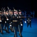A formal military drill or ceremonial parade is taking place on a darkened stage or indoor venue illuminated by blue lighting. Soldiers in dress uniforms are marching in formation, with the lead officer carrying a saber and a guidon bearer holding a unit pennant. The precision formation and formal attire indicate this is a ceremonial or honor guard performance.