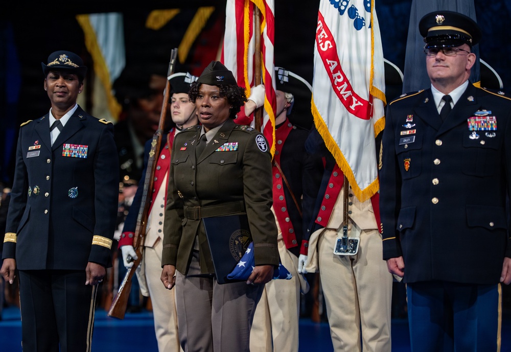 A formal U.S. Army military ceremony featuring uniformed personnel standing at attention on a stage or auditorium floor. A central figure in an olive drab Army Service Uniform holds a folded American flag and a blue folder, flanked by officers in dress blues and Army greens. Behind them, a color guard in historical Revolutionary War-era uniforms carries the U.S. flag and a United States Army flag.
