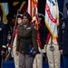 A formal U.S. Army military ceremony featuring uniformed personnel standing at attention on a stage or auditorium floor. A central figure in an olive drab Army Service Uniform holds a folded American flag and a blue folder, flanked by officers in dress blues and Army greens. Behind them, a color guard in historical Revolutionary War-era uniforms carries the U.S. flag and a United States Army flag.