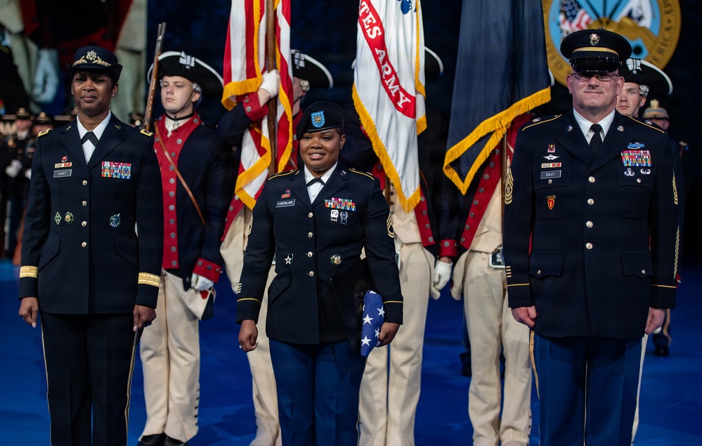 A formal U.S. Army military ceremony featuring three prominent uniformed soldiers standing at the forefront, with a color guard in Revolutionary War-era uniforms carrying flags including the U.S. flag and United States Army flag in the background. The central figure, a female Sergeant Major, holds a folded American flag, flanked by a female officer on the left and a male Sergeant Major on the right. The setting appears to be an indoor ceremonial venue with a blue floor.