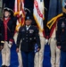 A formal U.S. Army military ceremony featuring three prominent uniformed soldiers standing at the forefront, with a color guard in Revolutionary War-era uniforms carrying flags including the U.S. flag and United States Army flag in the background. The central figure, a female Sergeant Major, holds a folded American flag, flanked by a female officer on the left and a male Sergeant Major on the right. The setting appears to be an indoor ceremonial venue with a blue floor.