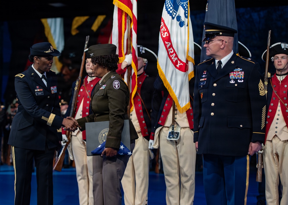 A formal U.S. Army military ceremony is taking place on a stage, featuring a handshake between two senior officers in dress uniforms. A color guard is present with the U.S. flag and U.S. Army flag, along with ceremonial soldiers dressed in Revolutionary War-era uniforms. The recipient appears to be holding a folded American flag and a presentation folder, suggesting a retirement or awards ceremony.