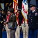 A formal U.S. Army military ceremony is taking place on a stage, featuring a handshake between two senior officers in dress uniforms. A color guard is present with the U.S. flag and U.S. Army flag, along with ceremonial soldiers dressed in Revolutionary War-era uniforms. The recipient appears to be holding a folded American flag and a presentation folder, suggesting a retirement or awards ceremony.