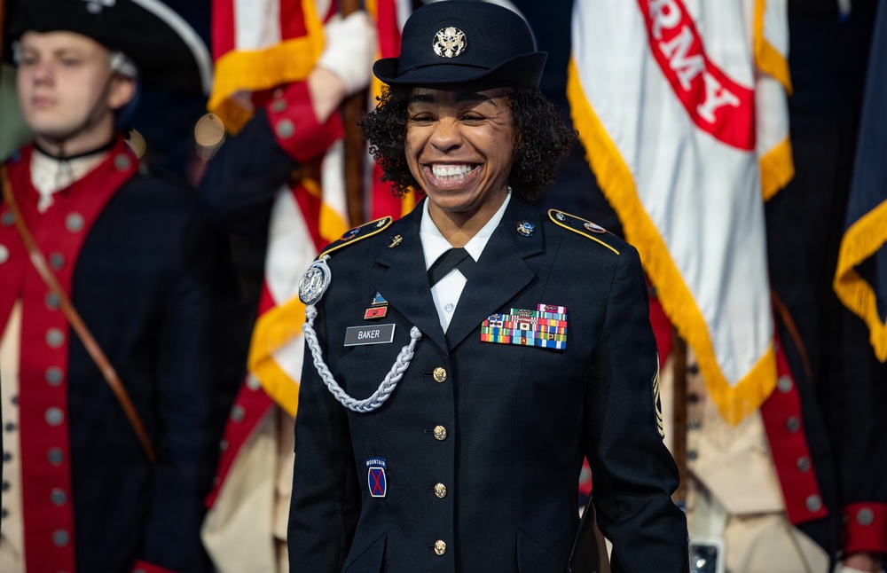 A U.S. Army soldier named Baker, wearing Army Service Uniform (ASU) with multiple service ribbons and a 10th Mountain Division patch, smiles broadly during what appears to be a formal military ceremony. She wears a distinctive silver aiguillette and a service cap with the U.S. Army crest. In the background, soldiers in Revolutionary War-era uniforms (Old Guard Fife and Drum Corps attire) carry flags including a visible U.S. Army flag.