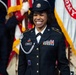 A U.S. Army soldier named Baker, wearing Army Service Uniform (ASU) with multiple service ribbons and a 10th Mountain Division patch, smiles broadly during what appears to be a formal military ceremony. She wears a distinctive silver aiguillette and a service cap with the U.S. Army crest. In the background, soldiers in Revolutionary War-era uniforms (Old Guard Fife and Drum Corps attire) carry flags including a visible U.S. Army flag.