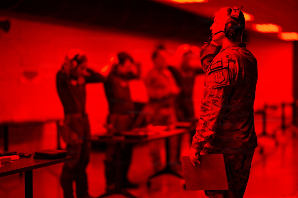 The image shows military personnel, identifiable by camouflage uniforms, in what appears to be an indoor training or briefing facility. The entire scene is bathed in a deep red light, creating a monochromatic atmosphere. The foreground figure is wearing hearing protection and holding a clipboard or folder, suggesting a supervisory or instructional role.