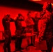 The image shows military personnel, identifiable by camouflage uniforms, in what appears to be an indoor training or briefing facility. The entire scene is bathed in a deep red light, creating a monochromatic atmosphere. The foreground figure is wearing hearing protection and holding a clipboard or folder, suggesting a supervisory or instructional role.