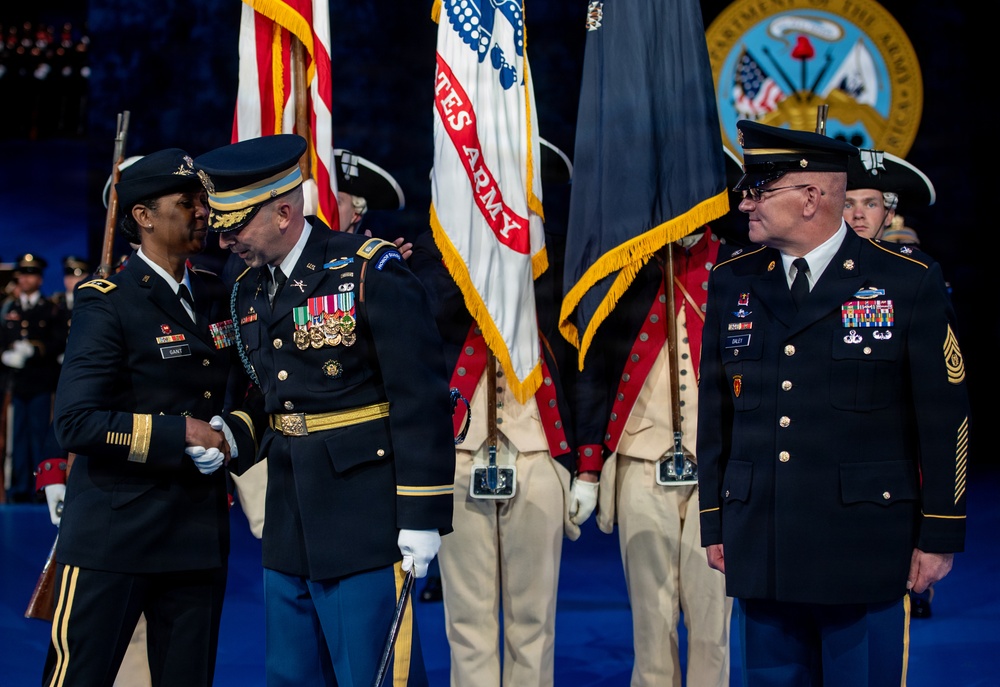 A formal U.S. Army military ceremony is taking place on a stage, featuring officers in dress uniforms engaged in what appears to be a handshake or award presentation. Multiple flags including the U.S. flag and U.S. Army flag are prominently displayed, along with soldiers in historical Revolutionary War-era uniforms serving as color guard. The Department of the Army seal is visible in the background, indicating a high-level official ceremony.
