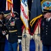 A formal U.S. Army military ceremony is taking place on a stage, featuring officers in dress uniforms engaged in what appears to be a handshake or award presentation. Multiple flags including the U.S. flag and U.S. Army flag are prominently displayed, along with soldiers in historical Revolutionary War-era uniforms serving as color guard. The Department of the Army seal is visible in the background, indicating a high-level official ceremony.