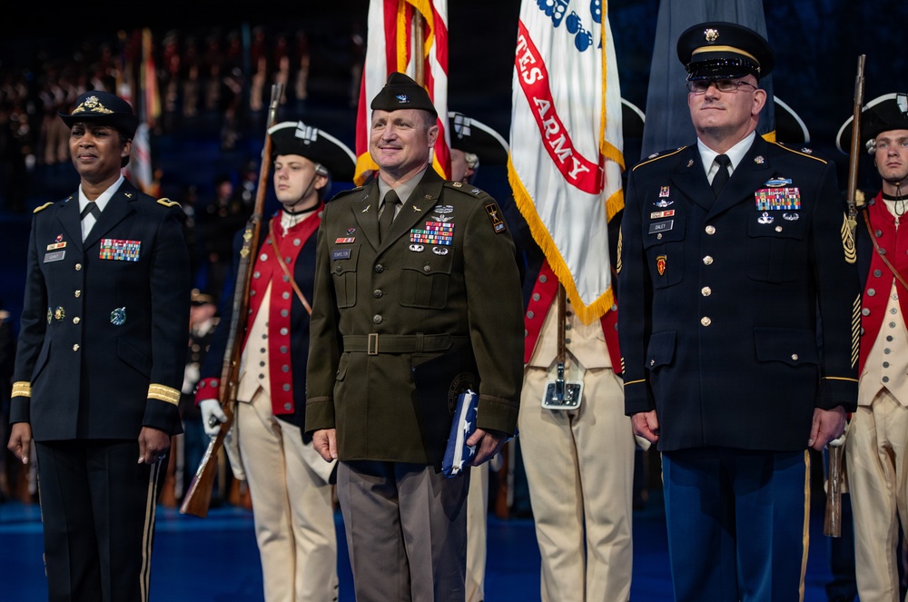 A formal U.S. Army military ceremony featuring personnel in multiple uniform types, including dress blues, Army greens, and colonial-era ceremonial uniforms. A central figure in Army green service uniform holds a folded flag, flanked by officers in dress blues and historical guard soldiers carrying rifles. A 'United States Army' flag is prominently displayed in the background.