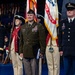 A formal U.S. Army military ceremony featuring personnel in multiple uniform types, including dress blues, Army greens, and colonial-era ceremonial uniforms. A central figure in Army green service uniform holds a folded flag, flanked by officers in dress blues and historical guard soldiers carrying rifles. A 'United States Army' flag is prominently displayed in the background.