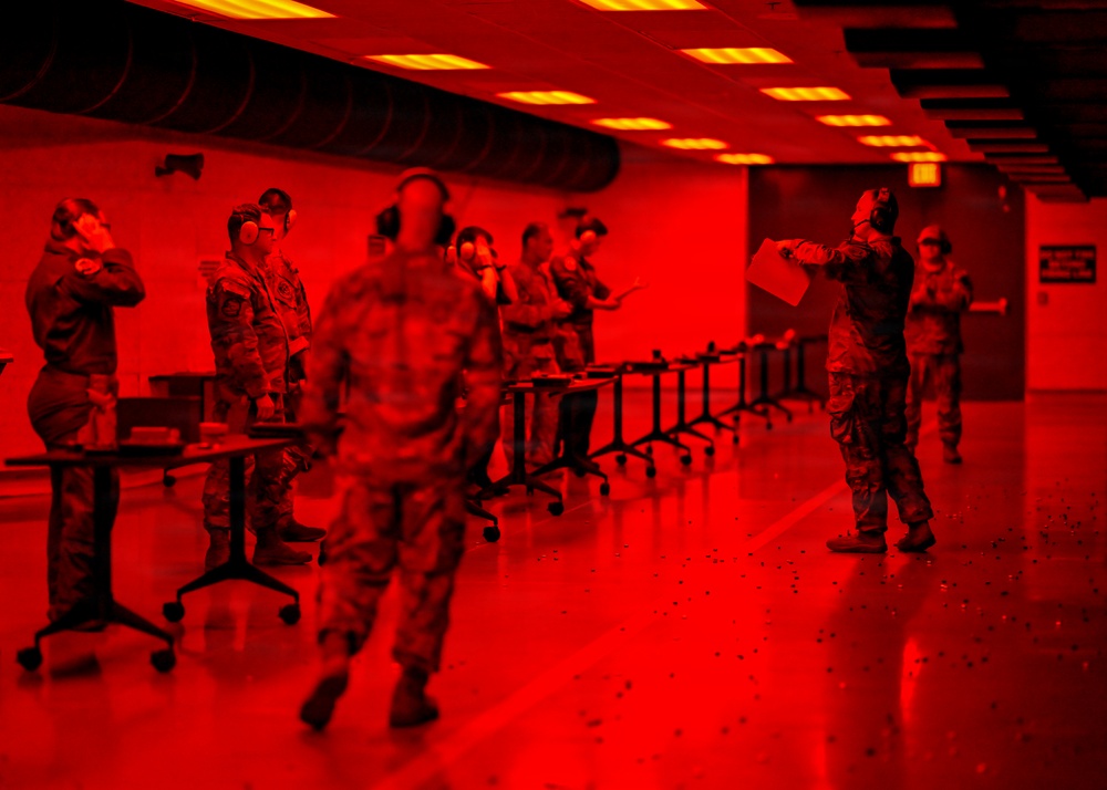 The image shows a group of military personnel in camouflage uniforms inside an indoor facility bathed in intense red lighting. Personnel are wearing hearing protection and appear to be engaged in a training or briefing exercise at a series of folding tables. One individual on the right appears to be an instructor or evaluator holding documents and wearing a gas mask.