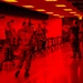 The image shows a group of military personnel in camouflage uniforms inside an indoor facility bathed in intense red lighting. Personnel are wearing hearing protection and appear to be engaged in a training or briefing exercise at a series of folding tables. One individual on the right appears to be an instructor or evaluator holding documents and wearing a gas mask.