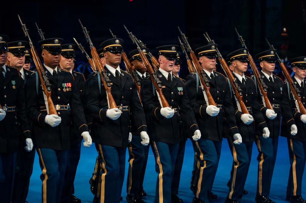 A precision military drill unit marches in formation during what appears to be an indoor ceremonial performance or tattoo event. The soldiers are dressed in formal dress uniforms and carry rifles with fixed bayonets at shoulder arms. The disciplined formation and synchronized movement suggest a highly rehearsed ceremonial drill routine.