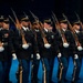 A precision military drill unit marches in formation during what appears to be an indoor ceremonial performance or tattoo event. The soldiers are dressed in formal dress uniforms and carry rifles with fixed bayonets at shoulder arms. The disciplined formation and synchronized movement suggest a highly rehearsed ceremonial drill routine.
