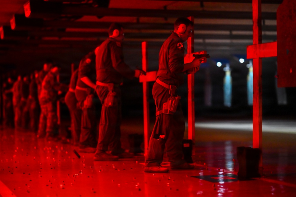 A group of individuals are participating in a firearms training exercise at an indoor shooting range illuminated by red lighting. Multiple shooters are lined up in shooting lanes, each positioned behind vertical dividers, holding handguns in a ready or inspection stance. The red ambient lighting and scattered brass casings on the floor suggest an active or recently completed live-fire training session.
