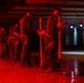 A group of individuals are participating in a firearms training exercise at an indoor shooting range illuminated by red lighting. Multiple shooters are lined up in shooting lanes, each positioned behind vertical dividers, holding handguns in a ready or inspection stance. The red ambient lighting and scattered brass casings on the floor suggest an active or recently completed live-fire training session.
