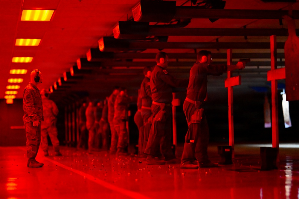 An indoor shooting range bathed in red lighting, showing a line of personnel in military/tactical attire conducting firearms qualification or training. Multiple shooters are positioned at firing lanes, with at least one supervisor or range safety officer observing from the left. The red ambient lighting and reflective floor create a dramatic, low-visibility atmosphere throughout the facility.