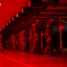 An indoor shooting range bathed in red lighting, showing a line of personnel in military/tactical attire conducting firearms qualification or training. Multiple shooters are positioned at firing lanes, with at least one supervisor or range safety officer observing from the left. The red ambient lighting and reflective floor create a dramatic, low-visibility atmosphere throughout the facility.