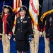 A formal U.S. Army military ceremony featuring three prominent service members standing at the forefront in dress uniforms. Behind them, a color guard in historical Revolutionary War-era uniforms carries flags including a U.S. Army banner. The center figure holds a folded American flag, suggesting a retirement, award, or commemorative ceremony.