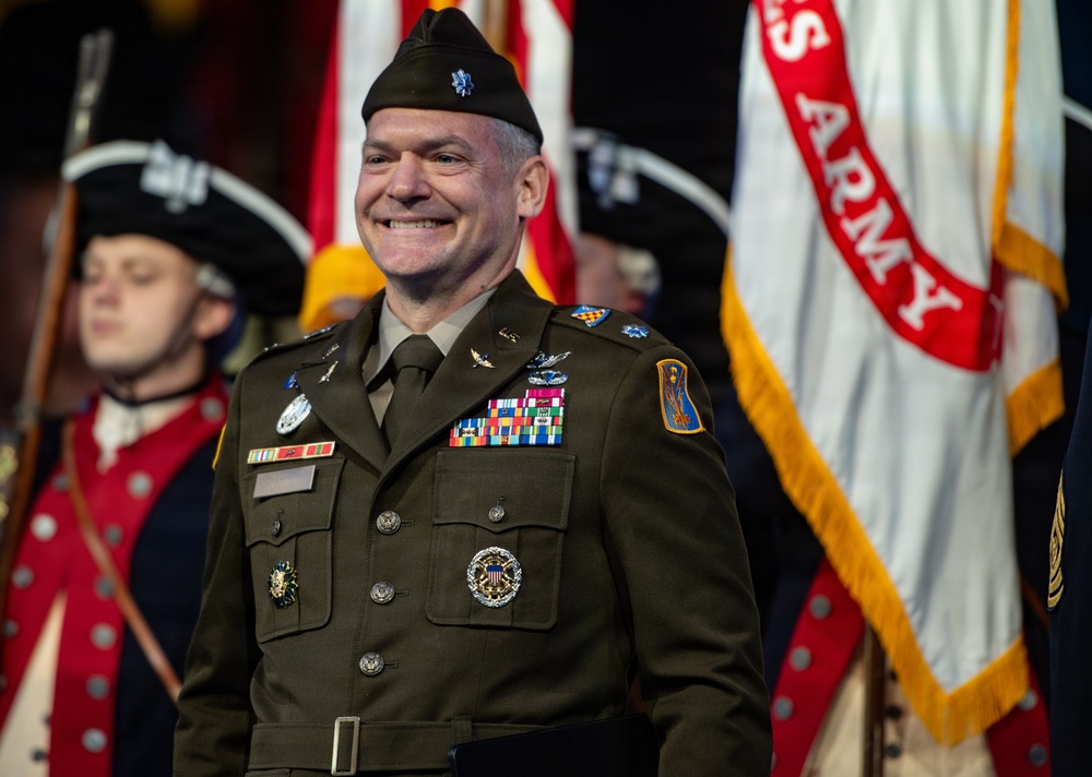 A smiling U.S. Army officer in dress green uniform stands at what appears to be a formal military ceremony. He is wearing a beret with a silver oak leaf cluster insignia, indicating the rank of Lieutenant Colonel, along with numerous service ribbons, badges, and medals. Behind him are ceremonial color guard soldiers in historical Revolutionary War-era uniforms and a U.S. Army flag.