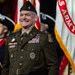 A smiling U.S. Army officer in dress green uniform stands at what appears to be a formal military ceremony. He is wearing a beret with a silver oak leaf cluster insignia, indicating the rank of Lieutenant Colonel, along with numerous service ribbons, badges, and medals. Behind him are ceremonial color guard soldiers in historical Revolutionary War-era uniforms and a U.S. Army flag.