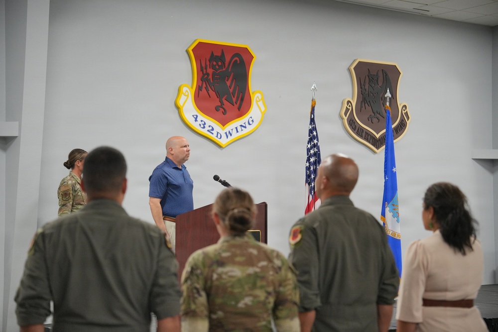 A formal ceremony or briefing is taking place at a U.S. Air Force installation, specifically associated with the 432d Wing and 432d Air Expeditionary Wing. A civilian man in a blue polo shirt is speaking at a wooden podium, while military personnel in flight suits and camouflage uniforms stand facing him. The setting features unit emblems/shields mounted on the wall and U.S. and Air Force flags flanking the speaker.