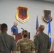 A formal ceremony or briefing is taking place at a U.S. Air Force installation, specifically associated with the 432d Wing and 432d Air Expeditionary Wing. A civilian man in a blue polo shirt is speaking at a wooden podium, while military personnel in flight suits and camouflage uniforms stand facing him. The setting features unit emblems/shields mounted on the wall and U.S. and Air Force flags flanking the speaker.