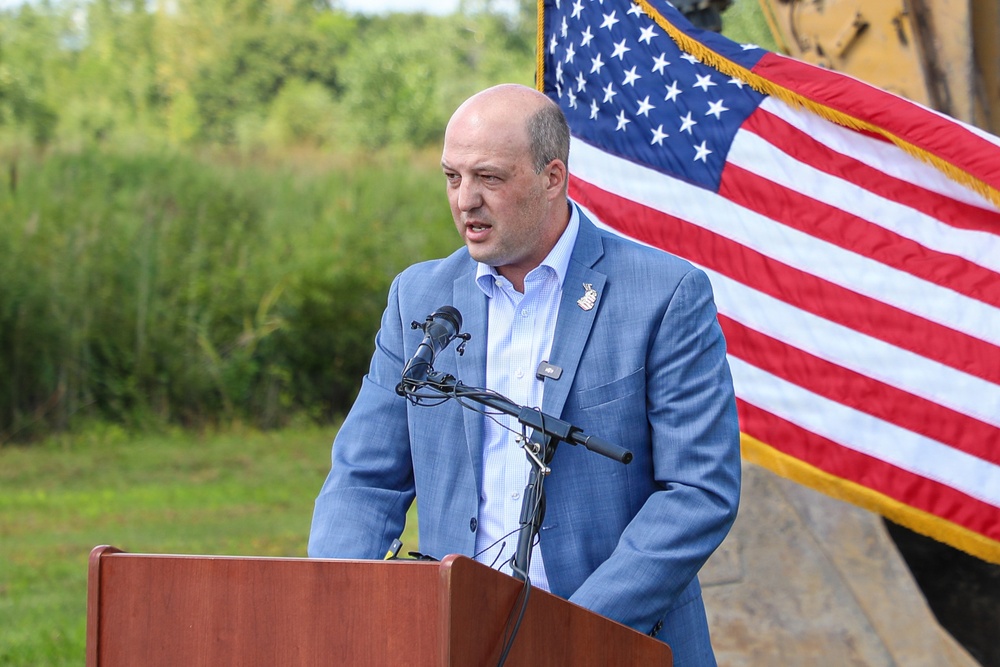 A man in a light blue suit jacket is speaking at a wooden podium equipped with a microphone setup at an outdoor event. An American flag with gold fringe is prominently displayed behind him to the right. The setting appears to be a public ceremony or official event held in a natural or semi-rural outdoor environment.