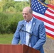 A man in a light blue suit jacket is speaking at a wooden podium equipped with a microphone setup at an outdoor event. An American flag with gold fringe is prominently displayed behind him to the right. The setting appears to be a public ceremony or official event held in a natural or semi-rural outdoor environment.