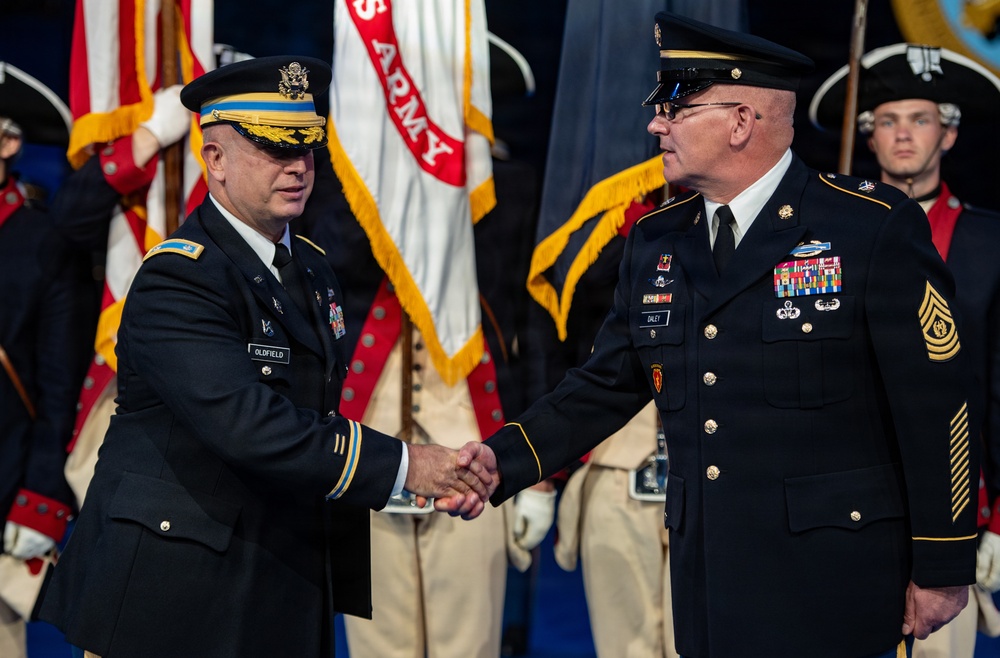 Two U.S. Army senior officers are shaking hands during a formal military ceremony, likely a change of command or retirement event. The officer on the left, identified by nameplate as 'OLDFIELD,' wears a general officer's cap with gold braid, while the officer on the right, identified as 'DALEY,' displays Sergeant Major rank insignia. A ceremonial color guard in historical Revolutionary War-era uniforms stands in the background alongside U.S. Army flags.