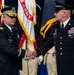Two U.S. Army senior officers are shaking hands during a formal military ceremony, likely a change of command or retirement event. The officer on the left, identified by nameplate as 'OLDFIELD,' wears a general officer's cap with gold braid, while the officer on the right, identified as 'DALEY,' displays Sergeant Major rank insignia. A ceremonial color guard in historical Revolutionary War-era uniforms stands in the background alongside U.S. Army flags.