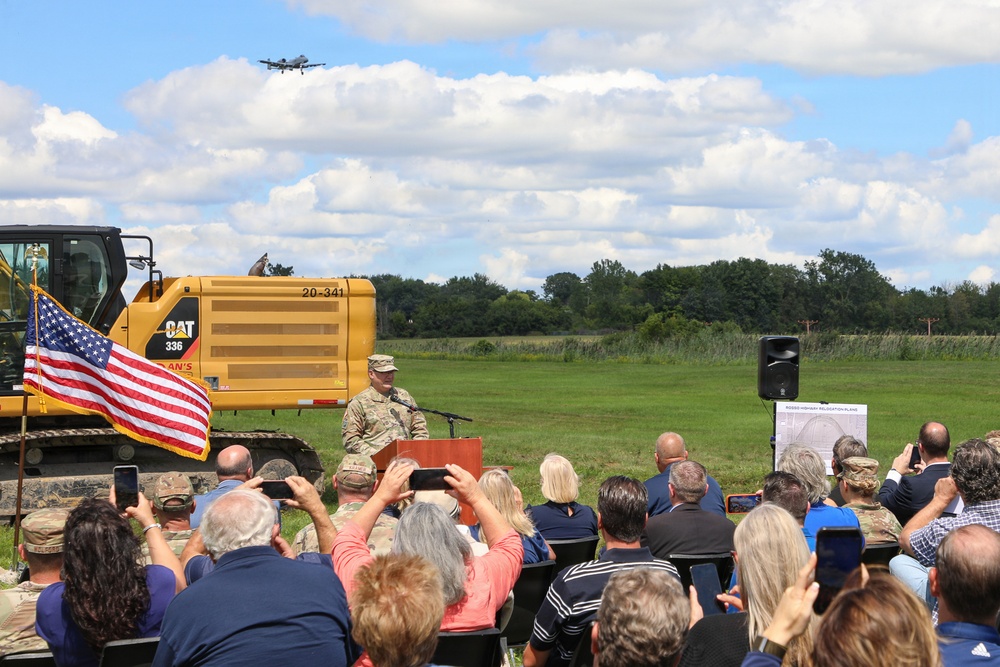 An outdoor groundbreaking ceremony is taking place at what appears to be a military installation or construction site. A uniformed military officer is speaking at a podium in front of a CAT 336 excavator, with an audience of civilians and military personnel seated and photographing the event. An aircraft is visible flying in the background sky.