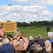 An outdoor groundbreaking ceremony is taking place at what appears to be a military installation or construction site. A uniformed military officer is speaking at a podium in front of a CAT 336 excavator, with an audience of civilians and military personnel seated and photographing the event. An aircraft is visible flying in the background sky.