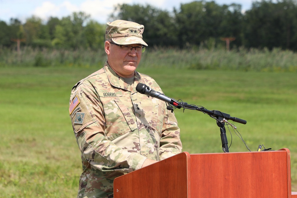 A U.S. Army officer with the name tape 'ROGERS' is speaking at a wooden podium with a microphone in an outdoor setting. The officer wears the Army Combat Uniform (OCP/MultiCam) and displays two stars on the chest, indicating the rank of Major General. The setting appears to be a formal military ceremony or event held on a grassy field.