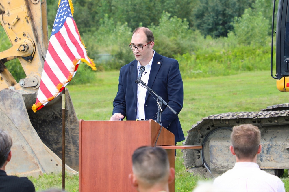 A man in a dark blazer is speaking at a podium with a microphone at what appears to be an outdoor groundbreaking ceremony. An American flag is displayed to his left, and yellow construction equipment (excavator) is visible on both sides of him. A small audience is visible in the foreground with their backs to the camera.