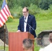 A man in a dark blazer is speaking at a podium with a microphone at what appears to be an outdoor groundbreaking ceremony. An American flag is displayed to his left, and yellow construction equipment (excavator) is visible on both sides of him. A small audience is visible in the foreground with their backs to the camera.