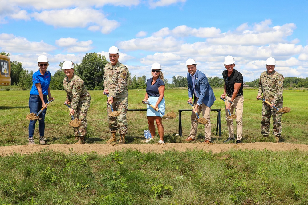 A groundbreaking ceremony is taking place outdoors on a grassy field, with seven participants holding gold-handled shovels with dirt. Three individuals are wearing military camouflage uniforms, while others are dressed in civilian attire, all wearing white hard hats. A CAT (Caterpillar) construction vehicle is partially visible in the background left.