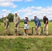 A groundbreaking ceremony is taking place outdoors on a grassy field, with seven participants holding gold-handled shovels with dirt. Three individuals are wearing military camouflage uniforms, while others are dressed in civilian attire, all wearing white hard hats. A CAT (Caterpillar) construction vehicle is partially visible in the background left.